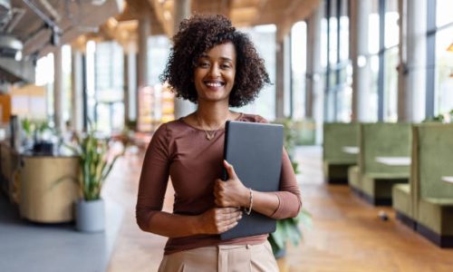 Portrait of happy African businesswoman holding digital tablet standing in the office. Female entrepreneur at her startup office looking at camera and smiling.