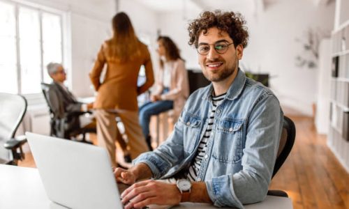 Portrait of young handsome businessman in the modern office, sitting at the desk, smiling, looking at camera