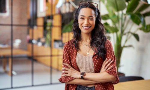 Portrait of smiling young multiethnic woman looking at camera with crossed arms. Successful latin business woman standing in modern office with copy space. Young university hispanic girl with pleasant smile and folded arms.