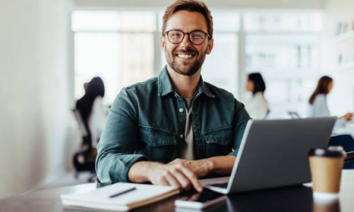 Portrait of a business man sitting in an office with his colleagues in the background. Happy business man working in a co-working office.